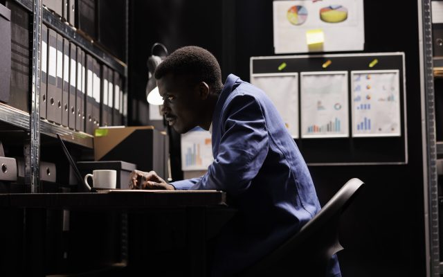 African american secretary looking at bookkeeping files, analyzing administrative documents late at night in storage room. Businessman discovering bureaucracy report, checking accountancy paperwork