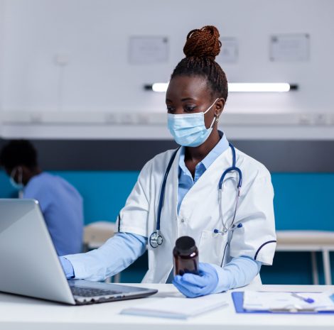 African american woman with doctor job holding bottle of medicine while using laptop on white desk in medical office. Black medic with uniform and stethoscope looking at digital screen