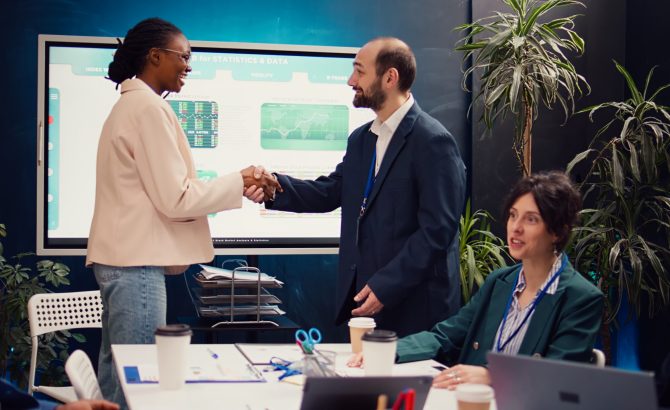 Business manager does handshake with a new employee during a briefing meeting, welcoming her to the team and presenting her to other staff members. Young man shaking hands with trainee. Camera B.